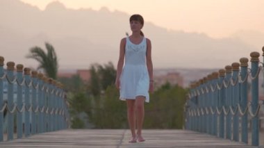 Akşamları sahilde uzun iskelede yürüyen beyaz elbiseli mutlu genç bir kadın. Tatiller ve seyahat konsepti