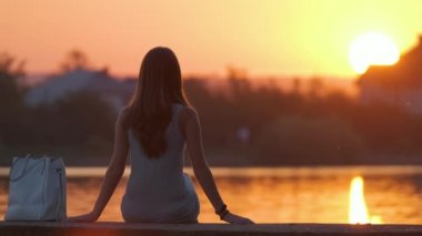 Sıcak bir akşamda göl kıyısında yalnız başına oturan yalnız bir kadın. Yalnızlık ve doğa konseptinde rahatlama.