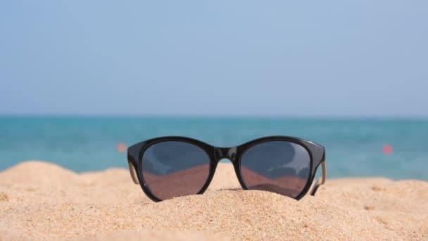 Primer plano de gafas de sol protectoras negras en la playa de arena en la costa tropical en un día cálido y soleado. Concepto vacaciones de verano — Vídeo de stock