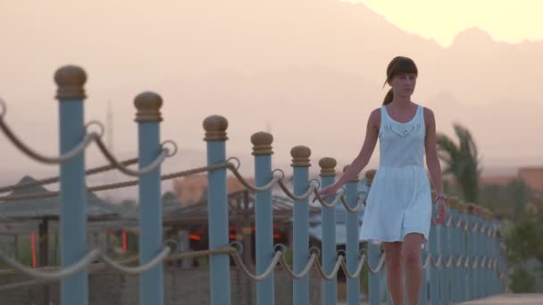 Fiatal, boldog nő fehér ruhában, hosszú mólón sétál este a tengerparton. Szabadságok és utazási koncepció — Stock videók