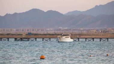Mavi deniz suyunun dalgalı yüzeyi ve sakin dalgalar üzerinde yüzen uzun rıhtımın sonundaki uzak yatıyla deniz manzarası.