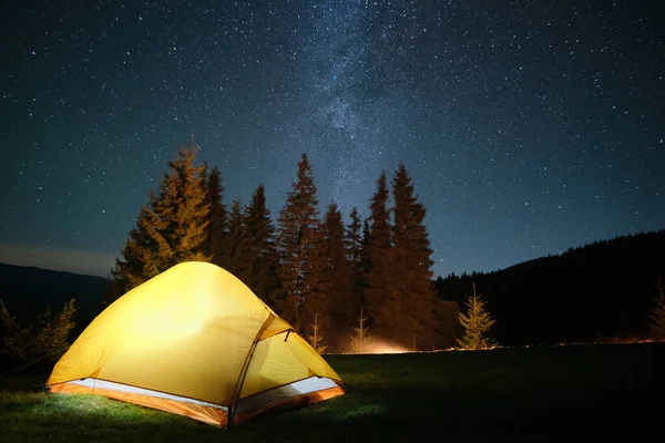 Bright illuminated tourist tent near glowing bonfire on camping site in dark mountains under night sky with sparkling stars. Active lifestyle concept — Stock Photo, Image