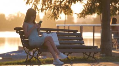 Parktaki bankta oturan genç bir kadın sıcak bir yaz akşamında cep telefonunu karıştırıyor. İletişim ve mobil bağlantı kavramı