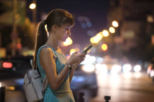Женщина с помощью телефона указывает пальцем на экран смартфона на фоне боке огней в ночной атмосфере освещенности города на вечерней разряженной улице. Онлайн-образ жизни с подключением к Интернету — стоковое фото