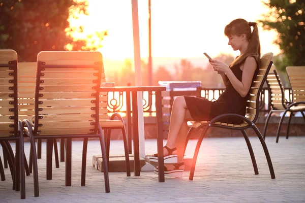 Молодая женщина, сидящая за столом уличного кафе, просматривает свой мобильный телефон под открытым небом теплым летним вечером. Концепция связи и мобильной связи. — стоковое фото