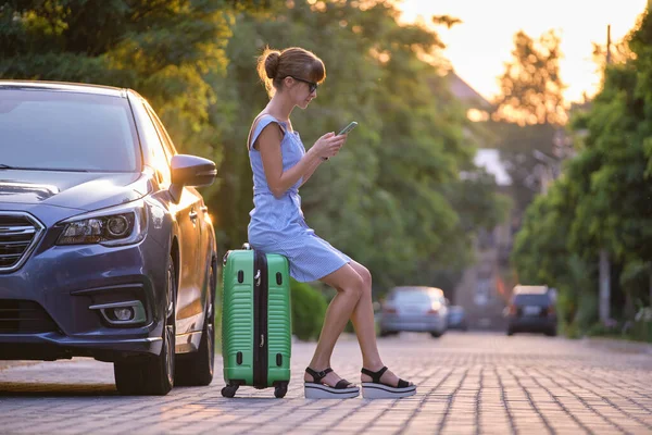 Mujer joven sentada en la maleta cerca de su vehículo hablando en su teléfono móvil en una calle de la ciudad en verano — Foto de Stock
