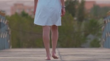 Akşamları sahilde uzun iskelede yürüyen beyaz elbiseli mutlu genç bir kadın. Tatiller ve seyahat konsepti