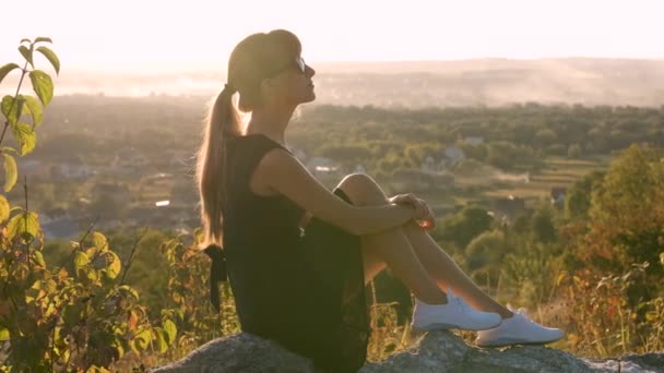 Mujer joven elegante en vestido corto negro y zapatos deportivos blancos sentados en una roca relajante al aire libre en la noche de verano. Dama de moda disfrutando de la cálida puesta de sol en la naturaleza — Vídeo de stock