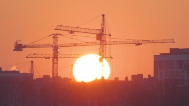 Gün batımında yüksek apartman binalarının inşaat sahasında büyük batan güneşli kule vinçlerinin koyu gölgesi. Gayrimenkul geliştirme