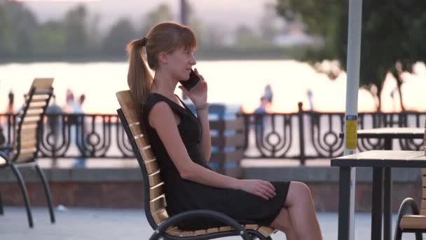 Giovane donna seduta al tavolo del caffè di strada a parlare sul suo cellulare all'aperto nella calda serata estiva. Concetto di comunicazione e connessione mobile — Video Stock