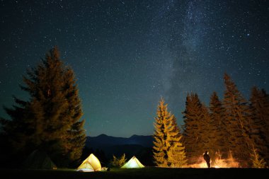 Yanlız yürüyüşçülerin silueti, karanlık dağlarda gece vakti gökyüzünün altında, aydınlık turist çadırlarının yanında şenlik ateşi yakmanın yanı sıra dinleniyor. Aktif yaşam tarzı ve açık hava yaşam konsepti