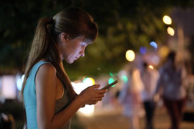 Gece atmosferik şehir aydınlatmasında arka planda akıllı telefon ekranını işaret eden bir kadın akşam vakti atmosferik şehir aydınlatmasında kullanılıyor. İnternet bağlantısı olan çevrimiçi yaşam biçimi