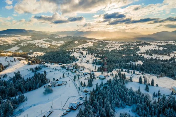 Aerial winter landscape with small village houses between snow covered forest in cold mountains in the evening — Stock Photo, Image