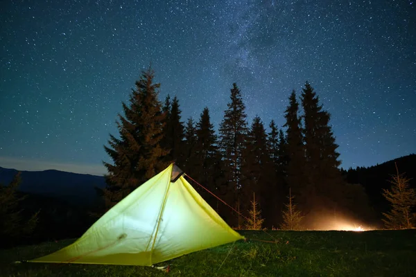 Bright illuminated tourist tent near glowing bonfire on camping site in dark mountains under night sky with sparkling stars. Active lifestyle concept — Stock Photo, Image