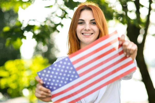 Portrait d'une jeune femme aux cheveux roux heureuse tenant le drapeau national des États-Unis dans ses mains debout à l'extérieur dans un parc d'été. Fille positive célébrant la fête de l'indépendance des États-Unis — Photo