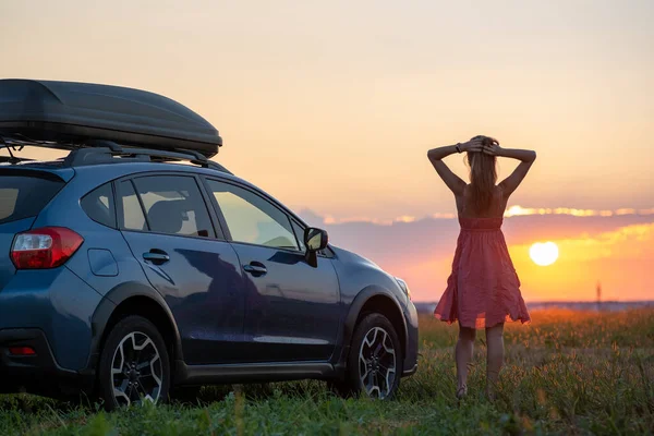 Silueta de conductora de pie cerca de su coche en el campo de hierba disfrutando de la vista de la puesta de sol brillante. Mujer joven que se relaja durante el viaje por carretera junto al vehículo SUV — Foto de Stock