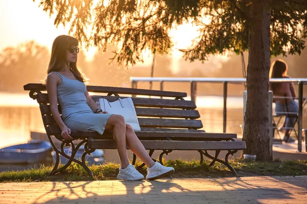 Молодая женщина в летнем платье отдыхает на скамейке у озера теплым вечером. Оздоровительный отдых от повседневной спешки — стоковое фото