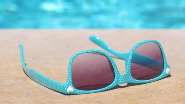 Gros plan de lunettes de soleil bleues sur le côté de la piscine par une chaude journée ensoleillée. Concept vacances d'été — Video