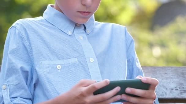 Ung pojke spelar spel på sin smartphone utomhus i sommarparken. Begreppet beroende av elektroniska prylar — Stockvideo