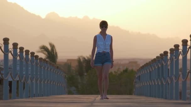 Trendig ung kvinna går en promenad på lång pir vid havet med siluett av bergstoppar på kvällen. Semester och resor koncept — Stockvideo