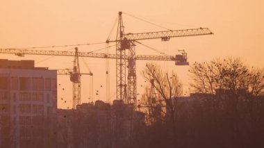 Gün batımında yüksek apartmanlı inşaat alanındaki vinçlerin koyu renkli silueti. Gayrimenkul geliştirme