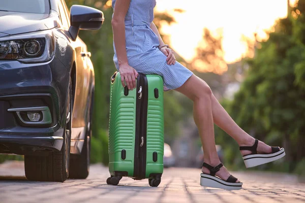 Nahaufnahme einer jungen Frau mit schlanken Beinen, die auf einer Koffertasche neben dem Auto ruht. Reise- und Urlaubskonzept — Stockfoto