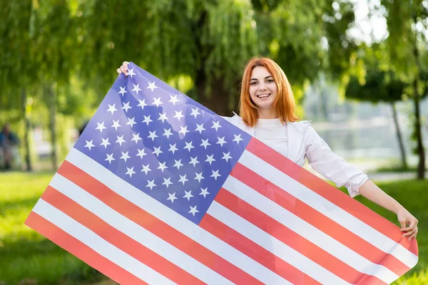 Szczęśliwa młoda rudowłosa kobieta pozująca z flagą USA stojącą na świeżym powietrzu w letnim parku. Pozytywna dziewczyna świętuje dzień niepodległości Stanów Zjednoczonych — Zdjęcie stockowe