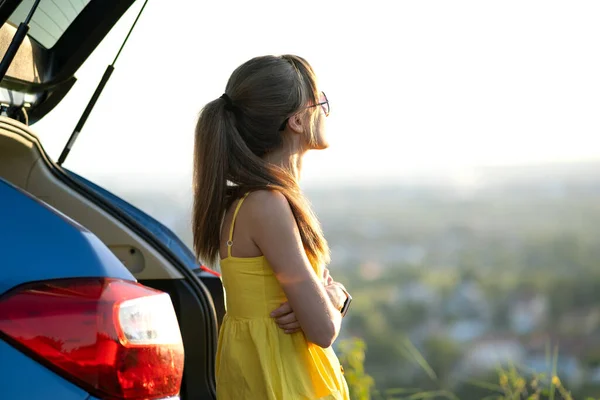 Mujer joven feliz en vestido amarillo de pie cerca de su vehículo mirando al atardecer vista de la naturaleza del verano. Concepto de viaje y vacaciones — Foto de Stock