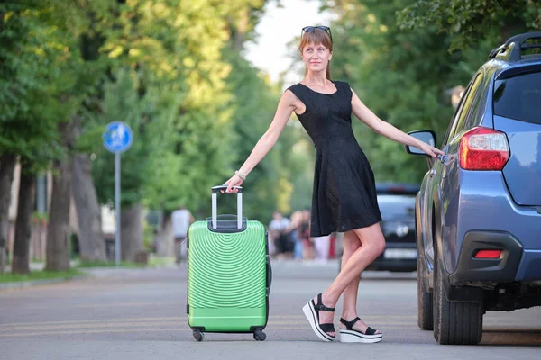 Glückliche junge Frau mit Koffertasche, die während einer Autofahrt neben ihrem Auto steht. Reise- und Urlaubskonzept — Stockfoto