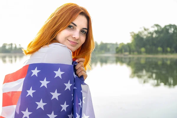 Portret szczęśliwej, uśmiechniętej rudej dziewczyny z flagą USA na ramionach. Pozytywna młoda kobieta świętująca dzień niepodległości Stanów Zjednoczonych — Zdjęcie stockowe