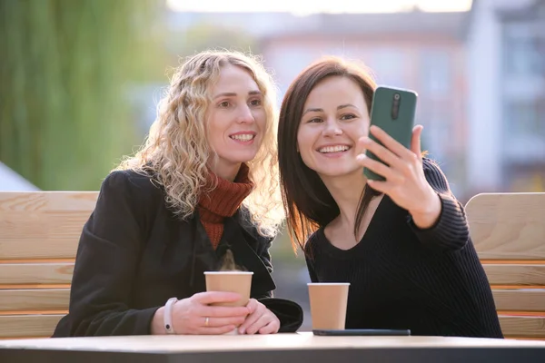 Две счастливые женщины делают селфи с мобильным телефоном, сидя в кафе на городской улице. Женщины-друзья наслаждаются временем вместе на открытом воздухе — стоковое фото