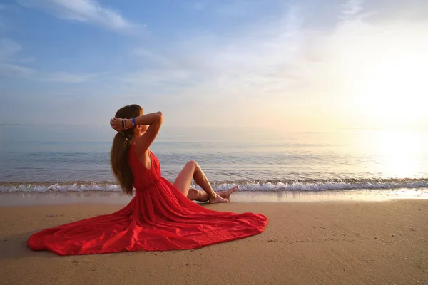 Young happy woman in red dress relaxing on sandy beach by seaside enjoying warm tropical morning — Stock Photo, Image