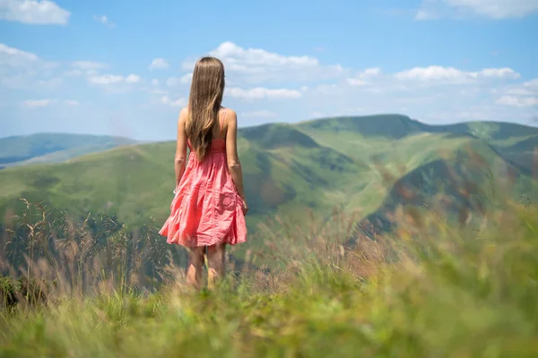 Genç, mutlu, kırmızı elbiseli bir gezgin yaz dağlarında rüzgarlı bir günde yeşil çimenli yamaçta dikilip doğanın tadını çıkarıyor.