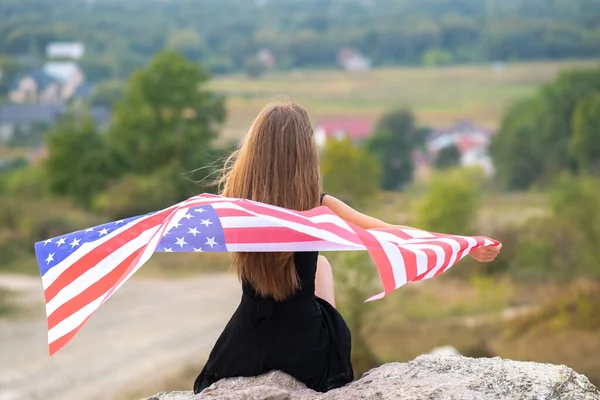 Jovem mulher americana bonita com cabelos longos segurando acenando no vento EUA bandeira em seus sholders descansando ao ar livre desfrutando de dia quente de verão — Fotografia de Stock