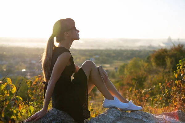 검은 짧은 옷을 입은 날씬 한 여자는 여름날해질 때 밖에서 휴식을 취하고 있는 바위 위에 앉아 있다. 자연계에서 따뜻 한 저녁을 즐기는 유행하는 여성 — 스톡 사진