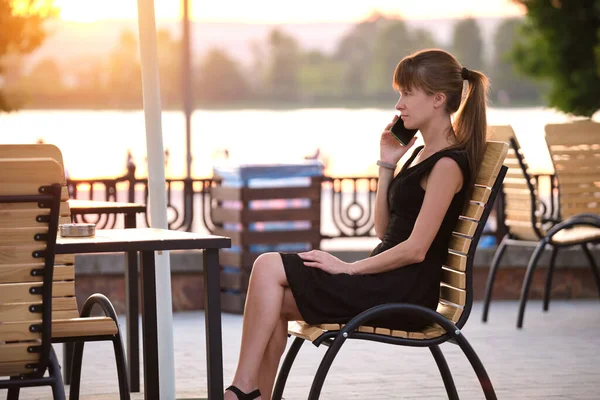 길거리 카페 탁자에 앉아 따뜻 한 여름날 저녁에 밖에서 핸드폰으로 이야기하는 젊은 여성 이 있었습니다. 의사소통 과 이동 통신 개념 — 스톡 사진