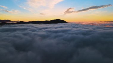 Ufuktaki uzak karanlık dağlarla beyaz yoğun bulutların üzerinde canlı gündoğumunun havadan görüntüsü.