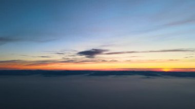 Beyaz, yoğun, sisli bulutların üzerindeki renkli günbatımının havadan görünüşü ufuktaki dağ tepelerinin uzak karanlık siluetleriyle kaplıdır.