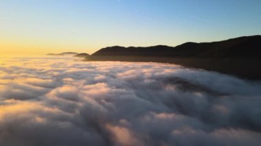 Ufuktaki dağ tepelerinin uzak karanlık siluetleri ile beyaz yoğun sis üzerinde renkli gün doğumunun havadan görüntüsü