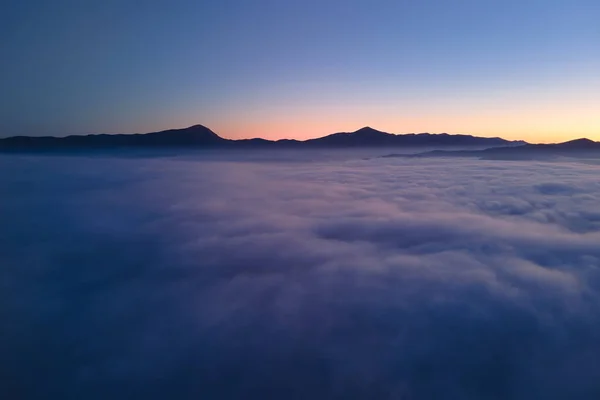 Vista aérea del colorido amanecer sobre la densa niebla blanca con siluetas oscuras distantes de colinas de montaña en el horizonte — Foto de Stock