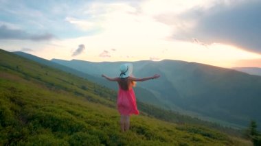 Kırmızılı genç kadın çimenli bir arazide rüzgarlı bir sonbahar akşamında ellerini kaldırarak doğanın tadını çıkarıyor.
