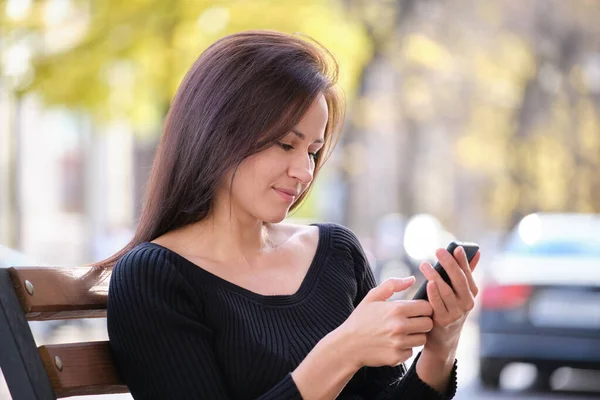 Молодая женщина, сидящая на уличной скамейке, просматривает свой мобильный телефон на улице в теплый летний день. Концепция связи и мобильной связи — стоковое фото