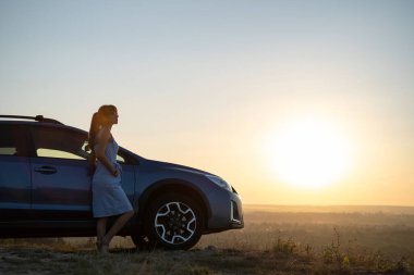 Mavi elbiseli mutlu genç kadın sürücü sıcak yaz akşamının tadını çıkarıyor arabasının yanında duruyor. Seyahat ve tatil kavramı