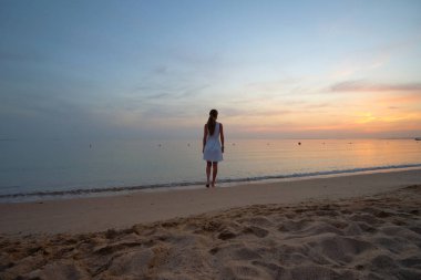 Yalnız genç bir kadın kumsalda duruyor, deniz kenarında sıcak tropikal bir akşamın tadını çıkarıyor.