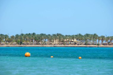 Deniz yüzeyinde yüzen sarı şamandıralar, kıyı şeridi manzaralı tropikal tatil beldesindeki derin sularda yüzme kısıtlamalarını işaret ediyor. İnsan hayatı güvenliği kavramı