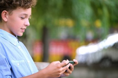 Yaz parkında akıllı telefonuyla oyun oynayan genç bir çocuk. Elektronik cihazlar konseptinden bağımlılık