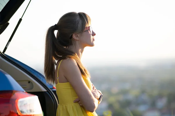 Conductora joven feliz en vestido amarillo apoyada en su coche disfrutando de un cálido día de verano. Concepto de viaje y vacaciones — Foto de Stock
