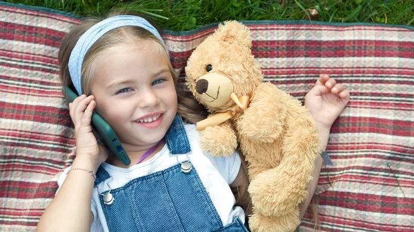 작고 행복 한 여자 아이가 잔디에 누워 테디 베어 와 휴대폰으로 얘기하는 모습 — 스톡 사진