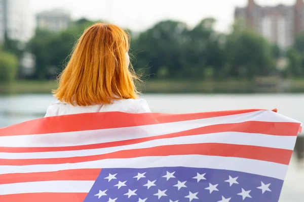 Portrait de fille aux cheveux rouges souriant heureux avec drapeau national des États-Unis sur les épaules. Une jeune femme célèbre la fête de l'indépendance des États-Unis — Photo