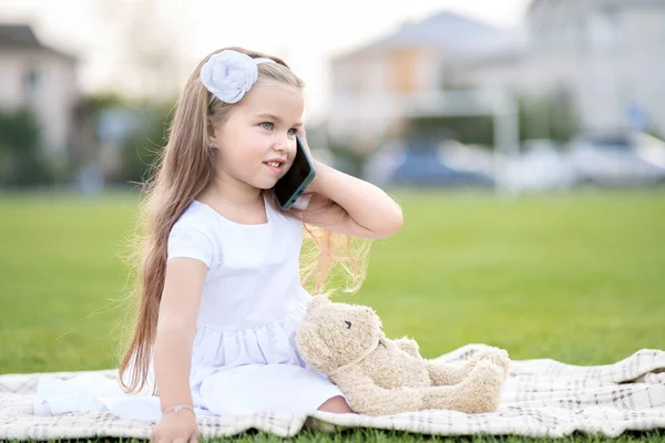 Söt liten flicka sitter i sommarparken tillsammans med sin nallebjörn leksak pratar på mobiltelefon ler glatt utomhus på sommaren — Stockfoto